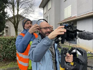 Thomas, gilet flo sur les épaules, place un casque audio sur les oreilles de Marc. Celui-ci a lesdeux mains prises par ses caméras. En fond un paysage pavillonaire.