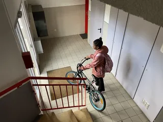Une jeune adolescent sort son vélo depuis son hall d'immeuble. Elle porte un manteau rose et un sac à dos.