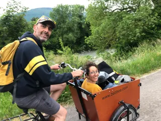 Cammile et Thomas voyage à Vélo argo. Camille est dans la caisse à l'avant. Thomas est au guidon. Tous deux regardent la caméra.