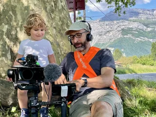 Marc, avec un gilet orange montre sont matériel de tournage à un enfant. Les deux sont sous un arbre. En fond une montagne.